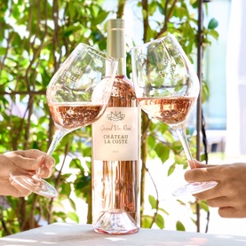 Chateau La Coste Grand Vin Ros&eacute; 1.5L bottle held between two clinking glasses of ros&eacute; wine, set against green foliage backdrop