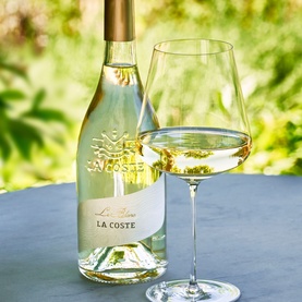 Chateau La Coste Le Blanc 500ml white wine bottle with gold label, styled alongside a filled crystal wine glass on an outdoor table.