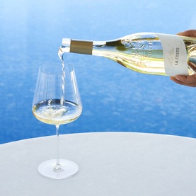 Chateau La Coste Le Blanc 75cl white wine being poured into a crystal wine glass, set against a bright blue background.