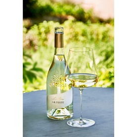 Chateau La Coste Le Blanc 75cl white wine bottle with gold tree motif, alongside a filled wine glass, styled outdoors on a grey surface.