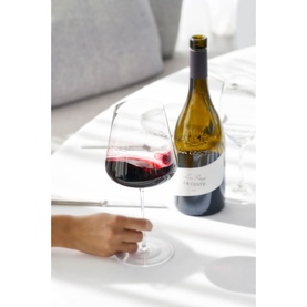 Chateau La Coste Le Rouge 75cl red wine bottle beside a filled large-bowled wine glass on a white table, hand reaching in