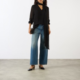 Model wearing House of Dagmar black wool blouse with choker neck and long tie detail, styled with wide-leg blue jeans and black mules.