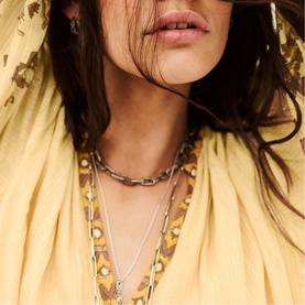 Model with Bash layered silver-tone chain necklaces, styled with a yellow floral print top and hat.