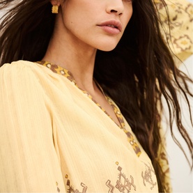 Model wearing Bash yellow embroidered top with V-neck, styled with gold earrings and beaded necklace, wide-brim hat visible