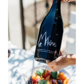 Chateau La Coste Maximas 750ml red wine bottle held by hand, dark glass with handwritten-style label, outdoor dining setting