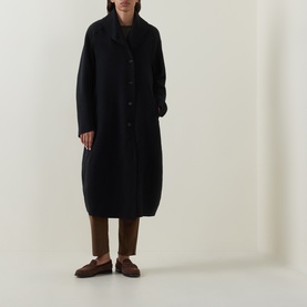 Model wearing Oska Fold-Down Wrap Coat in black with balloon hem and button placket, styled with brown trousers and loafers.