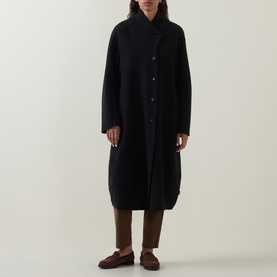 Model wearing Oska Fold-Down Wrap Coat in black; balloon hem, button placket, styled with brown trousers and loafers.