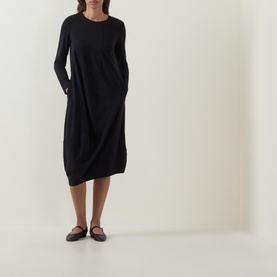 Model wearing Oska Solid Draped Dress in black; long sleeves, asymmetric draped midi hem, paired with black flat shoes.