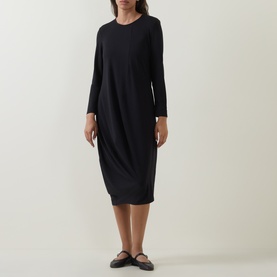 Model wearing Oska Solid Draped Dress in black; long sleeves, round neck, fluid midi length with asymmetric hem, styled with flat sandals.