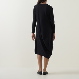 Model wearing Oska Solid Draped Dress in black, showing rear view with asymmetric hem and fluid drape; styled with black flat shoes.