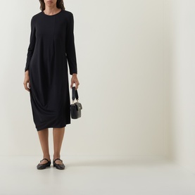 Model wearing Oska Solid Draped Dress in black, midi length with asymmetric hem and long sleeves, styled with black flat shoes and small bag.