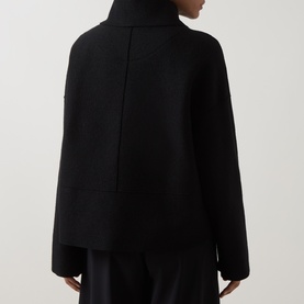 Model wearing Oska Fold-Down Wrap Jacket in black, back view showing dropped shoulders, exposed seam detail and long sleeves, styled with black