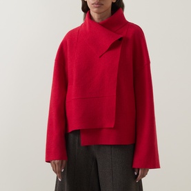Model wearing Oska scarlet wool jacket with asymmetric wrap front, high neck, and wide sleeves, styled with grey wide-leg trousers
