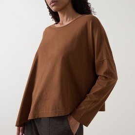 Model wearing Oska Asymmetric Long-Sleeved Top in Wood cotton, with round neckline, dropped shoulders and asymmetric hem, styled with grey trousers.
