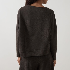 Model wearing Oska Asymmetric Knitted Sweater in Wood, rear view showing diagonal seam detail, dropped shoulders, styled with matching trousers.
