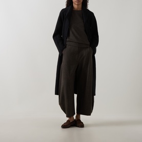 Model wearing Oska Asymmetric Knitted Sweater in Wood with wide-leg trousers, long black coat and brown loafers.