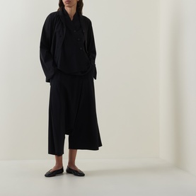 Model wearing Oska black wide-leg trousers with draped asymmetric hem, styled with a black oversized shirt and flat leather mules.