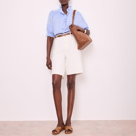 Model wearing Mint Velvet Blue Chambray Ruffle Blouse with cream shorts and tan belt, styled with brown woven bag and tan sandals.