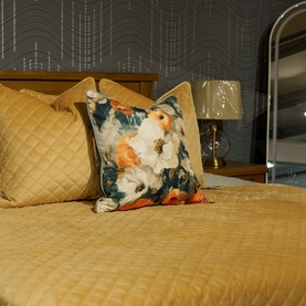 Fever + Hue Velvet Quilted Cushion in Caramel, styled on a bed with a floral print cushion and gold bedspread.