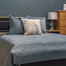 Fever + Hue Velvet Quilted Cushion in Gentle Blue, styled on a bed with matching bedspread and a floral feather print cushion.