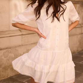 Model wearing white mini dress with pintuck detail, puff sleeves, button front, and tiered lace-trim hem