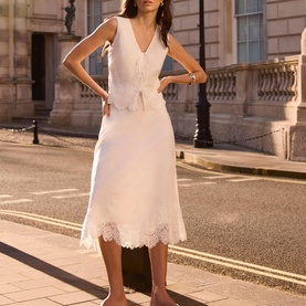 Model wearing white linen sleeveless V-neck vest top with button front, styled with matching white lace-hem midi skirt, shot on city street
