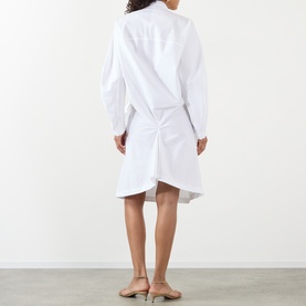 Model wearing Jacquemus white cotton shirt dress, shown from behind, with asymmetric ruched skirt detail and long sleeves, styled with nude sandals