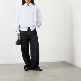 Model wearing Jacquemus grey cotton sweat top with white shirt underlayer, styled with black wide-leg jeans and black loafers, carrying mini black bag