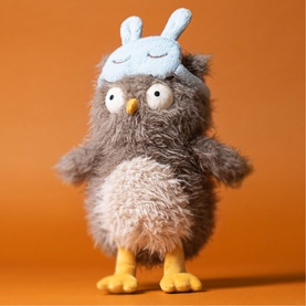 Jellycat grey fluffy owl soft toy with blue bunny ear sleep mask and yellow feet, standing upright against orange background