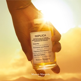Maison Margiela Replica EDT bottle held against a warm golden sunset, amber-toned glass flacon with label visible.