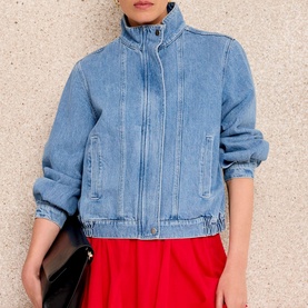 Model wearing light blue denim zip-up jacket with stand collar and front pockets, styled with red skirt and black clutch bag