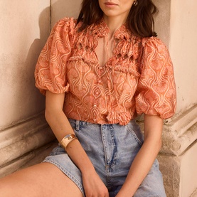 Model wearing Mint Velvet Orange Floral Ruffle Blouse with puff sleeves and ruffle bib, styled with light-wash denim shorts and a gold bracelet.