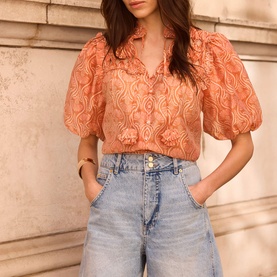 Model wearing Mint Velvet Orange Floral Ruffle Blouse with puff sleeves and ruffle bib, styled with light blue denim shorts.