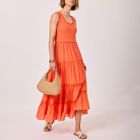Model wearing orange sleeveless tiered maxi dress, styled with strappy sandals and carrying a woven straw bag