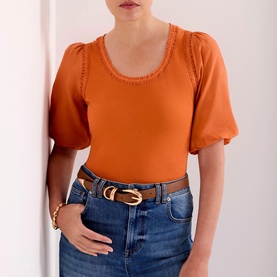 Model wearing orange puff-sleeve top with scoop neck and stitch trim detail, styled with blue jeans and tan leather belt