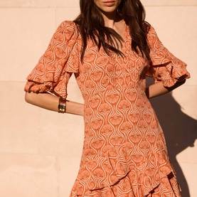 Model wearing orange abstract print mini dress with ruffle hem and flutter sleeves, styled with gold cuff bracelet