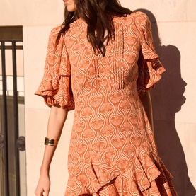 Model wearing orange and cream printed mini dress with puff sleeves, V-neck tie detail, pintuck front, and ruffle hem