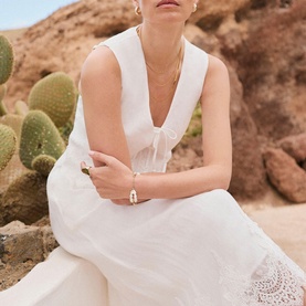 Model wearing Mint Velvet white sleeveless dress with deep V-neck and broderie anglaise hem, styled with gold jewellery, desert backdrop.