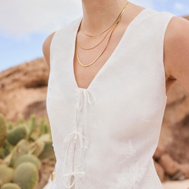 Model wearing Mint Velvet white sleeveless linen-look dress with tie-front detail, styled with layered gold chain necklaces