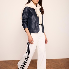 Model wearing navy faux leather jacket with cream shearling collar, styled with white wide-leg trousers featuring black side stripe detail.