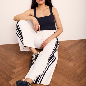 Model wearing white wide-leg trousers with navy stripe detail, styled with a navy square-neck tank top and navy trainers, seated on wooden floor