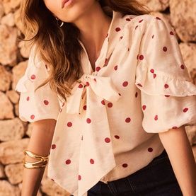 Model wearing Mint Velvet cream blouse with red polka dots, tie neck, puff sleeves and ruffle cuffs, styled with gold bangles.
