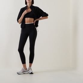 Model wearing Sweaty Betty black jacket, styled with black sports bra, matching leggings and grey trainers