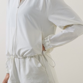 Model wearing Sweaty Betty jacket in Lily White, showing zip-front fastening, elasticated waist with drawstring, and ruched cuffs.