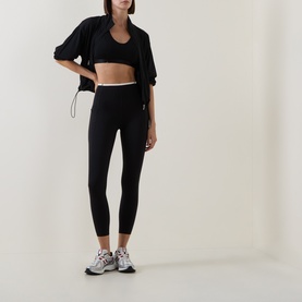 Model wearing Sweaty Betty black leggings with white waistband trim, styled with black sports bra, zip jacket and white trainers