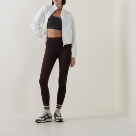 Model wearing Sweaty Betty Espresso Brown leggings, styled with a white zip-up jacket, black sports bra, and trainers.
