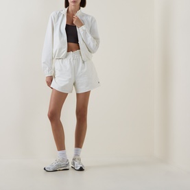 Model wearing Sweaty Betty Lily White shorts, styled with matching white zip jacket, black crop top and white trainers.