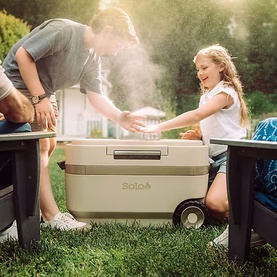 Solo Stove Windchill 47 Cooler & Fan in sandy beige, used outdoors on grass by adults and a child at a garden gathering