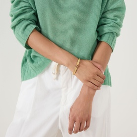 Model with Coach Logo Textured Hinge Bangle Bracelet in shiny gold, worn on wrist. Styled with green knit jumper and white trousers.