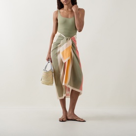 Model wearing Eres Croquant Printed Sarong in multi, block stripe print with self-tie. Styled with green swimsuit, carrying woven bag.